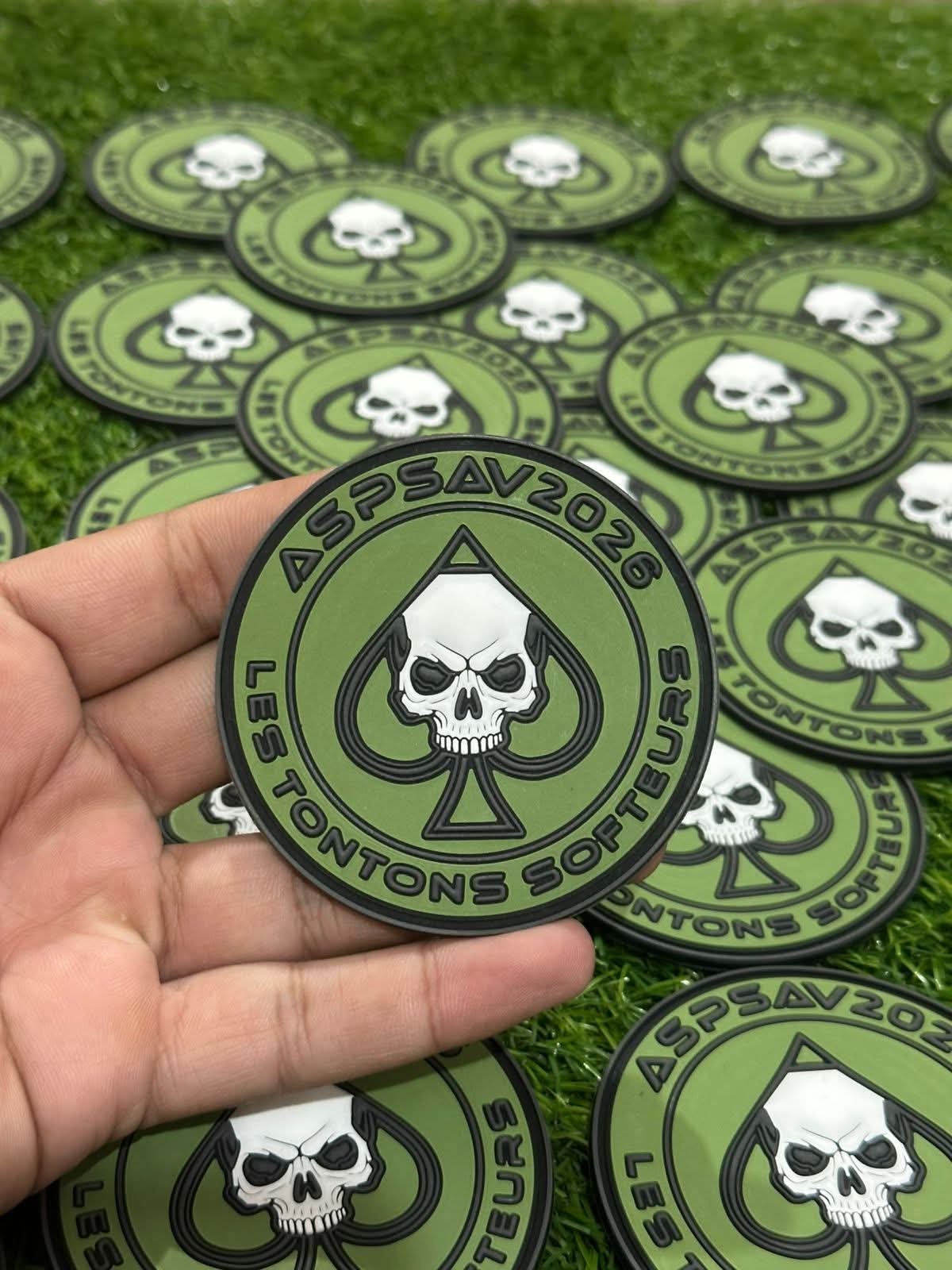 ASPSAV 2026 tactical green PVC morale patch with white skull inside a black spade emblem, displayed in hand with multiple matching patches in the background. Custom Tactical Rubber Patch