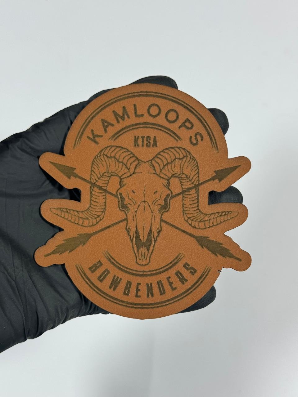 Brown laser engraved Kamloops Bowbenders leatherette patch featuring a ram skull with crossed arrows and custom text, designed for hunting, outdoor, and tactical gear. Custom Leatherette Ram Skull Patch